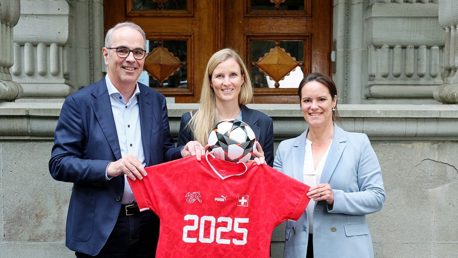 Stadpräsident Beat Züsli, Leevke Stutz und Regierungsrätin Michaela Tschuor halten ein Trikot mit der Aufschrift "2025" und ein Fussball in die Höhe.